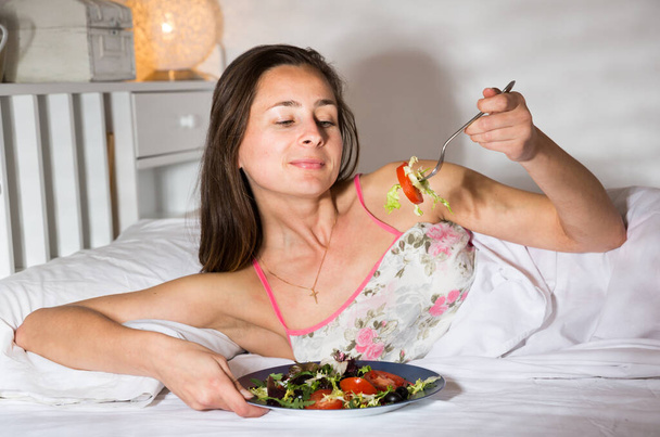 Woman in nightgown coat eats salad in bed - Photo, Image