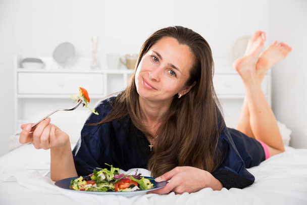Young pretty woman eating vegetable salad from plato in bed - Foto, imagen