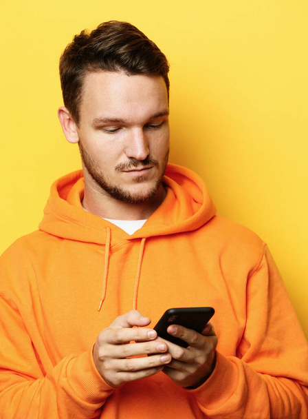 young handsome man holding mobile phone and making photo of himself while standing against yellow background - Valokuva, kuva