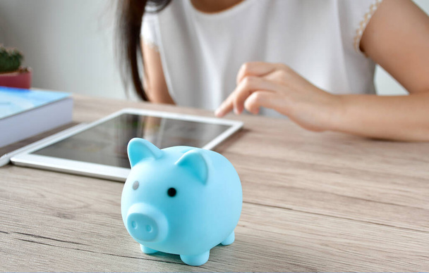 A woman calculating the savings on the iPad. There is also a blue piggy bank on the wooden table to save money and save household income on the tablet. - Photo, Image