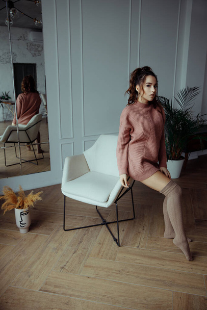 A woman in a warm pink sweater and knee socks is sitting on the back of a chair. - Фото, изображение