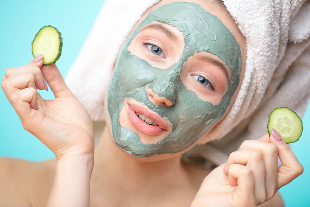 Funny young girl in clay mask holding slice cucumber for eyes treatment - Photo, Image