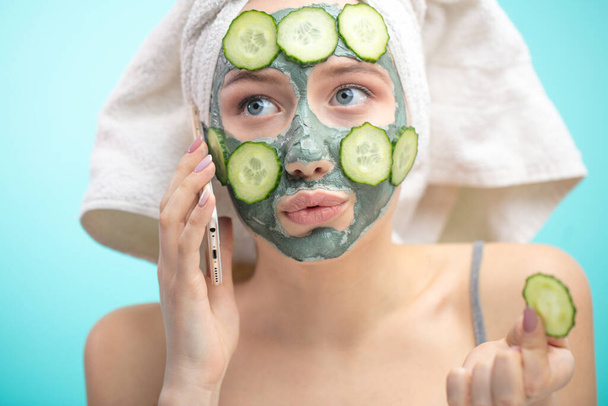 Woman with towel on head and face mask enriching with cucumbers via smartphone - Photo, image