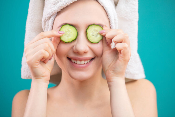 Woman with a towel on her head having fun, covering eyes with cucumber slices. - Фото, зображення