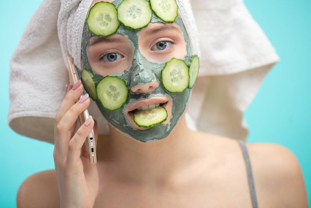 Woman with towel on head and face mask enriching with cucumbers via smartphone - Fotografie, Obrázek