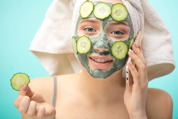 Woman with towel on head and face mask enriching with cucumbers via smartphone - Foto, immagini