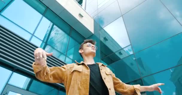 Closeup elegante cara atraente adolescente homem usa jaqueta na moda e óculos de sol, danças em movimentos modernos de rua, gira à mão, se move flexível de forma flexível, se diverte ao ar livre perto de vidro edifício urbano - Filmagem, Vídeo