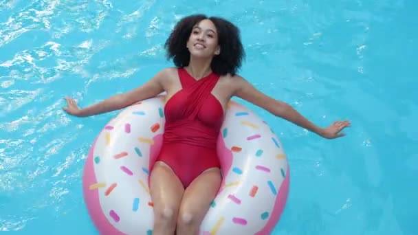Afrikaans model Amerikaanse vrouw zit liggend in opblaasbare zwemring donut, roeien snijdt het blauwe water in het zwembad met haar handen, tans, glimlacht kijken naar camera 's, sluit haar ogen, ontspant, bovenaanzicht - Video