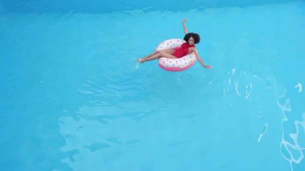 A young swimmer teenage girl sits in inflatable life-saving circle inblue clean cool water in sea pool, restores strength, makes waves on surface, moves her arms like butterfly rowing, distant plan - Footage, Video