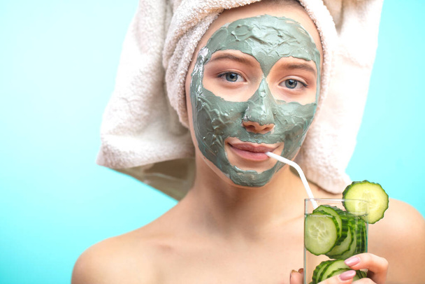 woman with cosmetic clay mask drinking cucucmber water isolated at studio blue - Фото, зображення