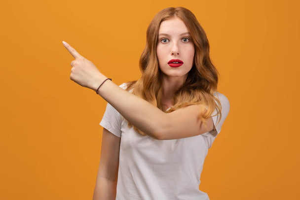 Pretty girl with wavy redhead, wearing white t-shirt, surprised and pointing finger to the side, shows copy space for your information. Isolated on yellow background - Photo, Image