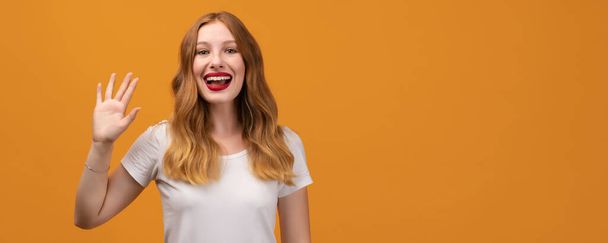 Hello. Portrait of adorable friendly woman with wavy redhead, wearing white t-shirt, standing waving hand, looking at camera with engaging toothy smile. Copy space for your text - Photo, Image