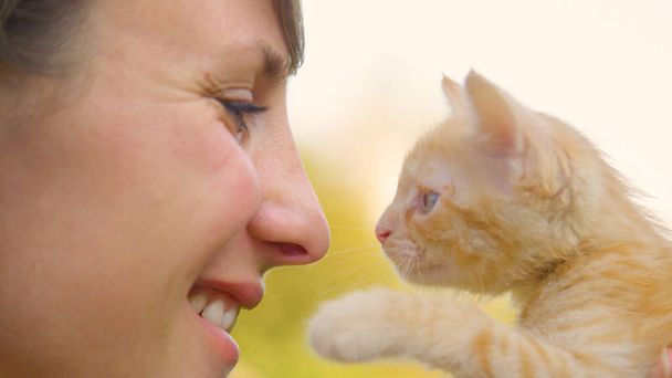 CLOSE UP: Junge Frau lächelt, als sie eine süße orangefarbene Katze vor ihr Gesicht hebt. - Foto, Bild