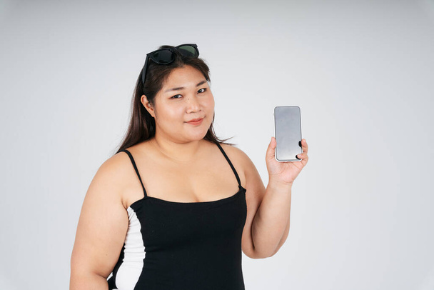 Portrait de femme potelée asiatique en robe noire montrant l'écran du smartphone sur fond blanc pour simuler ou copier l'espace. - Photo, image