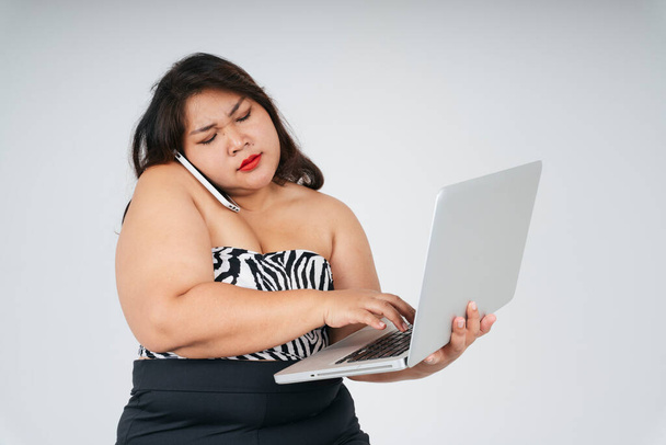 Busy asian chubby woman holding laptop and talking on the phone with serious looking on white background. - Foto, Imagem