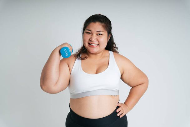 Asian chubby woman in white sport bra exercise with blue dumbbell over white background. - Fotó, kép