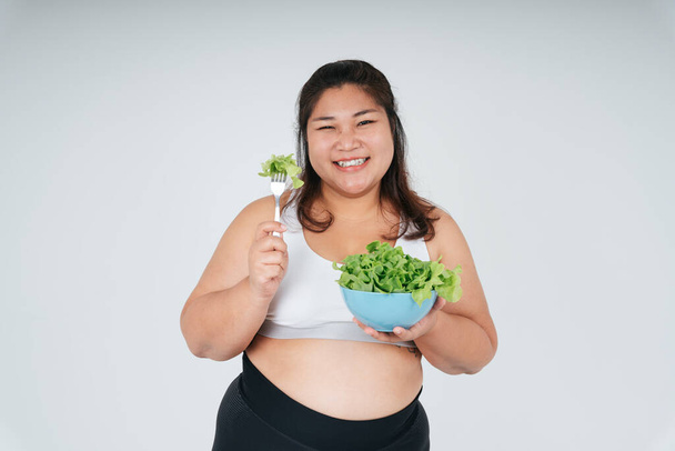 Cheerful happy asian chubby woman in white sport bra enjoy eating salad bowl over white background. - Foto, Imagen