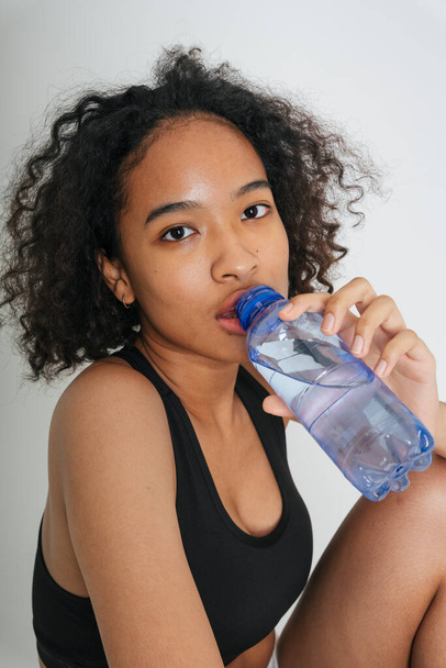 Смешанная раса африканка и тайка пьют воду из бутылки на белом фоне. - Фото, изображение