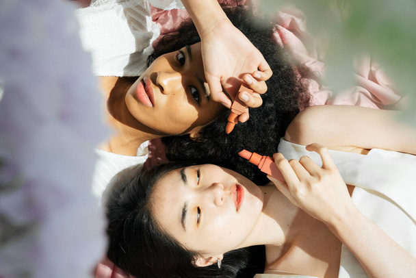 Top view - Asian and african women in white shirt holdin lipstick laying on pink cloth. - Фото, зображення