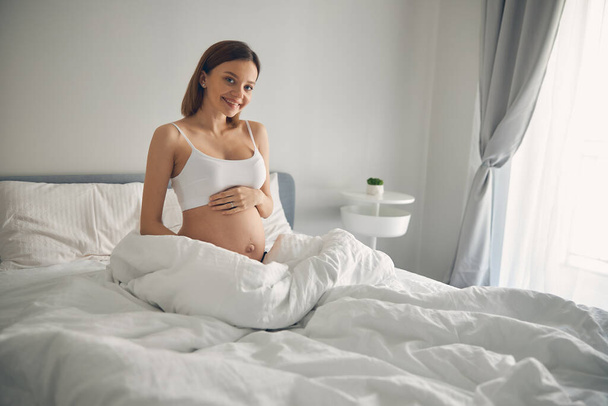 Smiling young lady caressing her belly in the white apartment - Foto, immagini