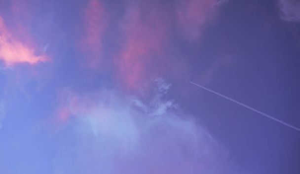 L'aereo vola attraverso il cielo e lascia dietro di sé un pennacchio bianco. Bellissimo tramonto estivo cielo blu con nuvole rosa. Il sole illumina il cielo in modo che diventi rosa e arancione. - Foto, immagini
