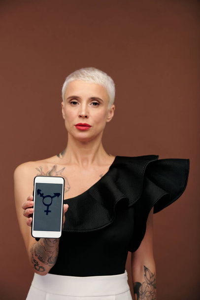 Moderne elegante Frau mit Tätowierungen an Armen und Brust und kurzen blonden Haaren, Smartphone mit Transgender-Symbol auf dem Bildschirm - Foto, Bild