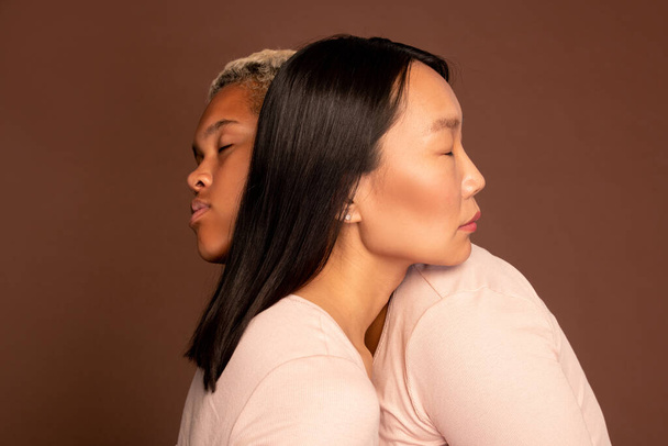 Jóvenes hembras asiáticas y afroamericanas con los ojos cerrados abrazándose y manteniendo la cabeza sobre los hombros - Foto, imagen