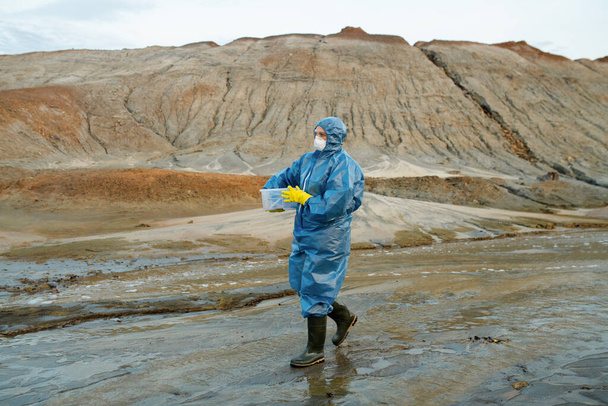 Contemporary female scientist in protective coveralls, respirator, rubber gloves and boots carrying plastic container against hills and sky - Photo, Image