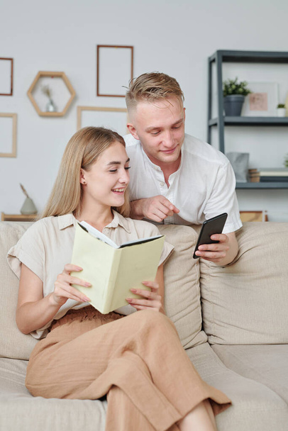 Young man in white shirt showing his wife with book photo of new model of home appliance in smartphone while surfing in online shop - Фото, изображение