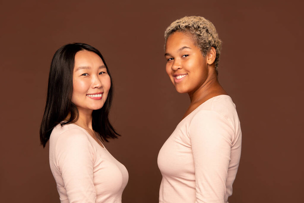 Dos jóvenes hembras alegres con sonrisas dentales mirando a la cámara mientras están de pie uno frente al otro contra el fondo marrón - Foto, Imagen