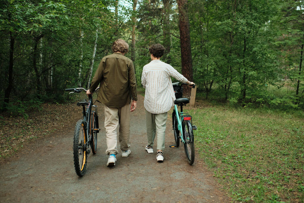 Вид сзади на молодую пару в повседневной одежде, разговаривающую друг с другом при движении по лесной дорожке или дороге между зелеными деревьями - Фото, изображение