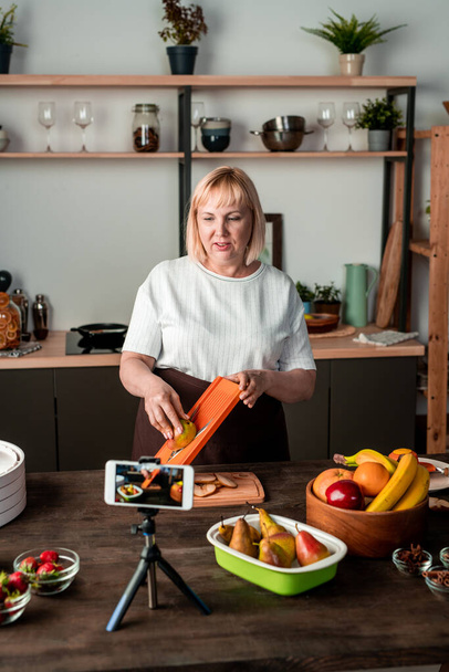 Случайная домохозяйка режет свежие груши на доске, чтобы высушить их в бытовой электросушилке, стоя у кухонного стола - Фото, изображение