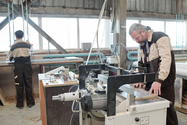 Сірошерстий зрілий працівник заводу фіксує дошку на робочій поверхні, збираючись обробити її на промисловій машині в робочому середовищі
 - Фото, зображення