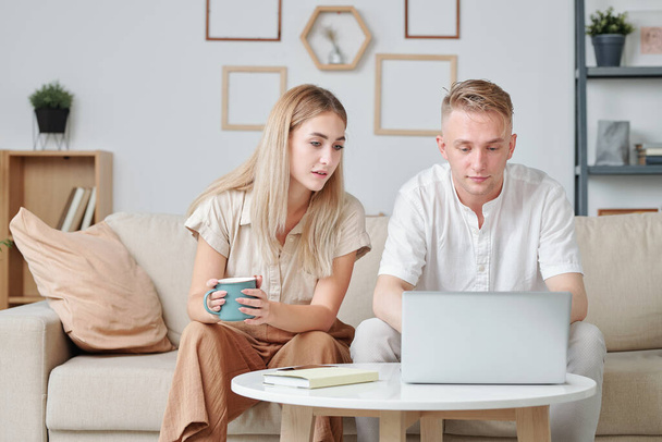 Mujer joven con taza de bebida sentada en el sofá junto a su marido con el surf portátil en la tienda en línea, mientras que la elección de nuevo gadget - Foto, Imagen