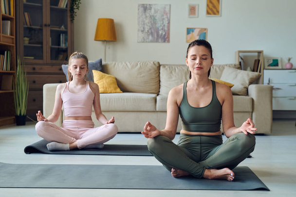 Розслаблена брюнетка жінка і її дочка-підліток в трекових костюмах, сидячи на килимках в позі лотоса, беручи курс йоги для початківців вдома
 - Фото, зображення