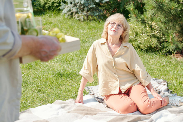 Unrecognizable senior man bringing food and lemonade to his wife waiting for him to start picnic on lawn - Foto, immagini