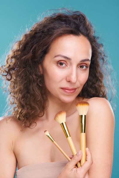 Linda jovem fêmea com pele saudável segurando três escovas cosméticas profissionais enquanto vai aplicar blush, pó e sombra para os olhos - Foto, Imagem