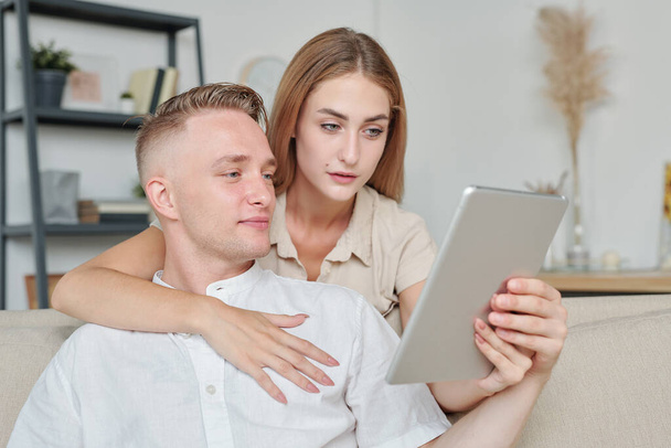 Uczuciowa młoda kobieta z tabletem obejmującym jej męża, pokazując mu ładne ubrania, które chciałaby kupić w sklepie internetowym - Zdjęcie, obraz