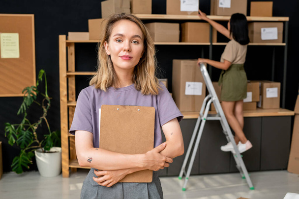 Jovem trabalhadora loira confiante de sala de armazenamento com área de transferência em pé na frente da câmera contra seu colega na escada rolante - Foto, Imagem