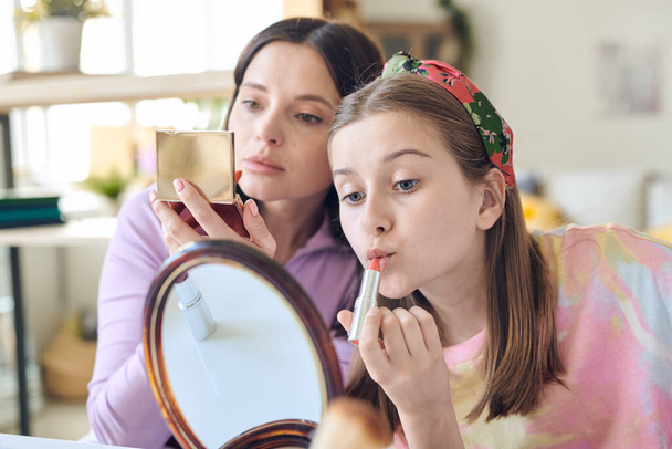 Schattig tienermeisje met lang blond haar aanbrengen van roze lippenstift en kijken in spiegel op de achtergrond van haar moeder met open compacte poeder - Foto, afbeelding