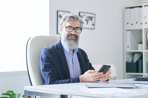 Retrato de hombre de negocios maduro con estilo con barba en la cara sentado en el escritorio de la oficina sosteniendo el teléfono inteligente mirando a la cámara - Foto, imagen