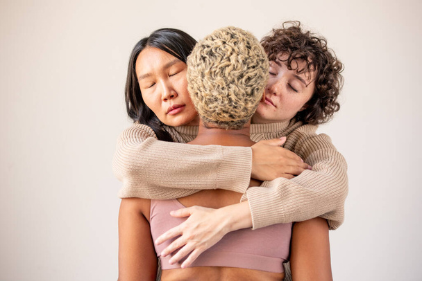 Two young females of Caucasian and Asian ethnicities in one sweater embracing African woman with short dyed blond hair in isolation - Photo, Image