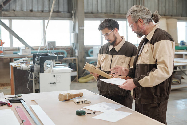 Пожилой профессиональный инженер, объясняющий молодому стажеру эскиз деревянной детали на встрече за большим столом в цехе завода - Фото, изображение