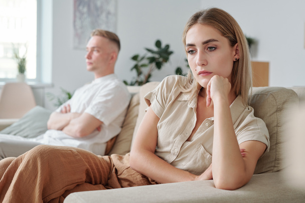 jong verdrietig vrouw en haar beledigde echtgenoot op de achtergrond zitten op de bank in de woonkamer en houden stilte na of tijdens ruzie - Foto, afbeelding