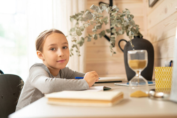 Jolie écolière en pull gris assise à table et tenant un stylo sur un carnet ouvert tout en vous regardant et en faisant des devoirs - Photo, image