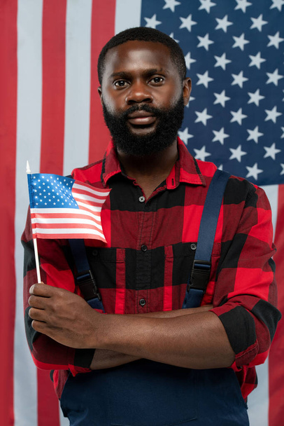 Young bearded African-american worker in flannel and overalls holding flag while standing against stars-and-stripes background - Zdjęcie, obraz