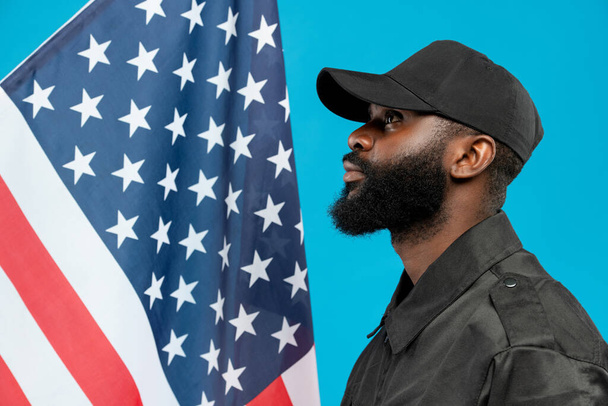 Молодий афро-американський чоловічий захист в чорній формі і шапка стоять перед камерою проти зірко-смугастого прапора - Фото, зображення