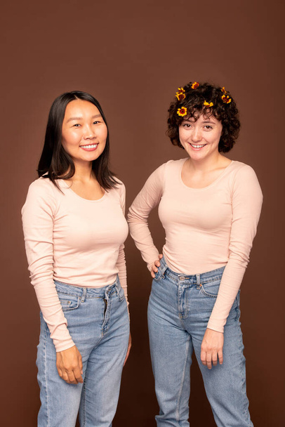 Duas jovens mulheres interculturais felizes em pulôveres brancos e jeans azuis de pé perto um do outro na frente da câmera e olhando para você - Foto, Imagem