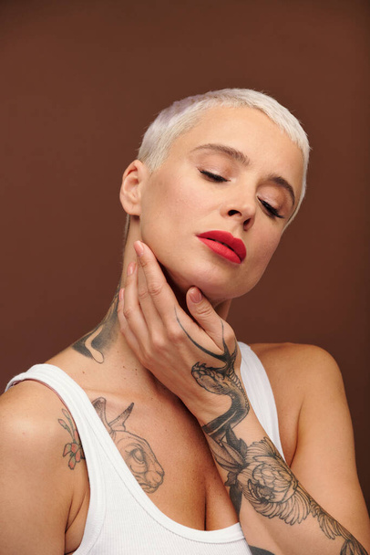 Mature blond female with tattooes on arm touching her face by hand and keeping her eyes closed while standing in front of camera in isolation - Foto, afbeelding