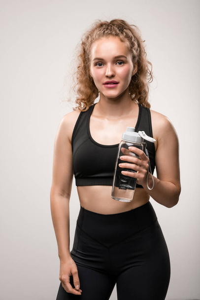 Junge, gut gelaunte Frau in schwarzem Trainingsanzug hält Plastikflasche mit reinem Wasser in der Pause zur Erfrischung nach dem Sport - Foto, Bild